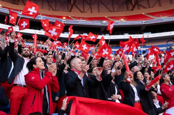 The audience in a stadium waves small Swiss flags and applauds.