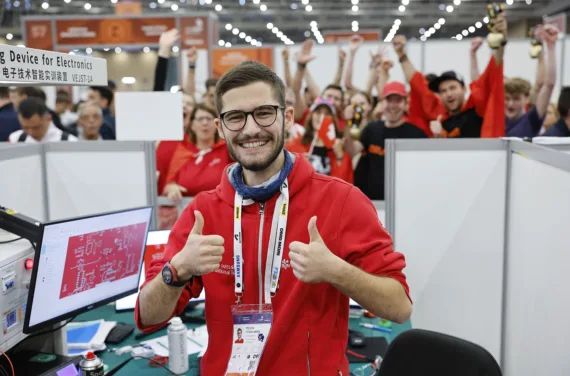 A young man in a red jumper smiles and gives a thumbs up in front of an enthusiastic crowd holding Swiss flags.