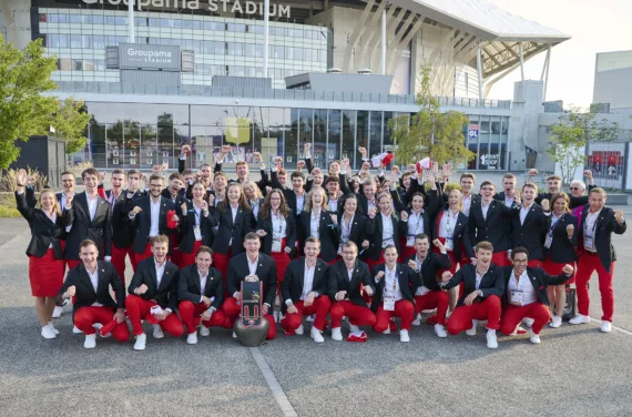 L'équipe suisse en pantalons rouges, blazers sombres et chemises blanches pose devant un stade.