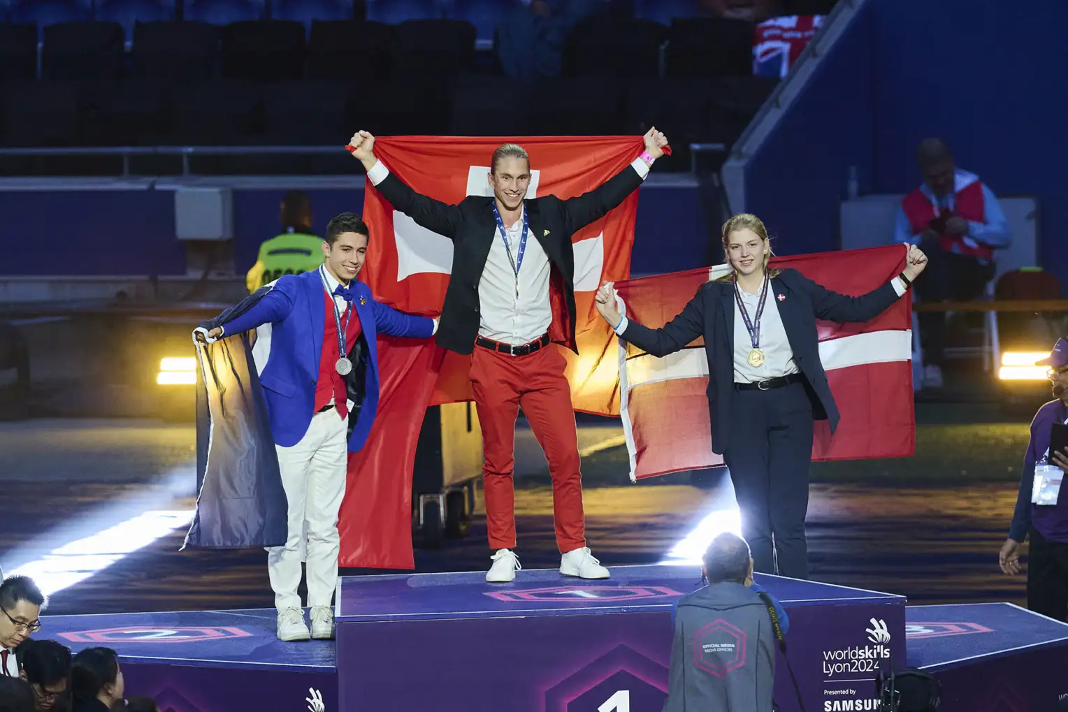 Trois jeunes gens se tiennent sur un podium et tiennent leurs drapeaux nationaux derrière eux: une Autrichienne à la troisième place, un Français à la deuxième place, et un Suisse à la première place.