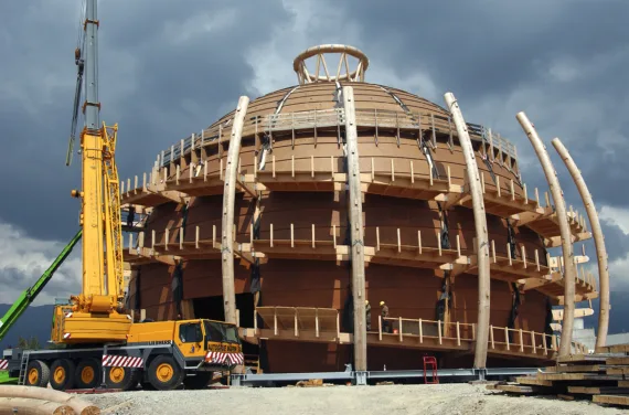 Photographie d'un dôme en bois brun-rouge en construction avec une grue jaune sur sa gauche.