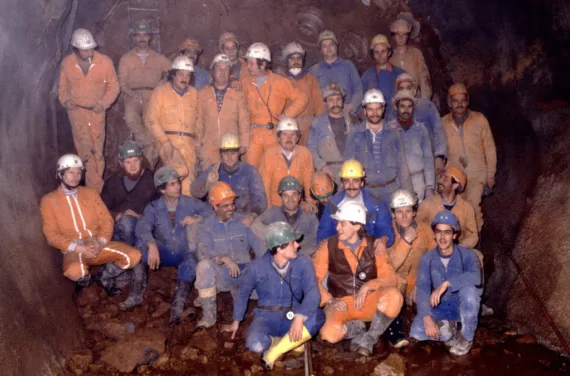 A construction crew in orange and blue overalls wearing safety hats poses for a group photo in a tunnel.