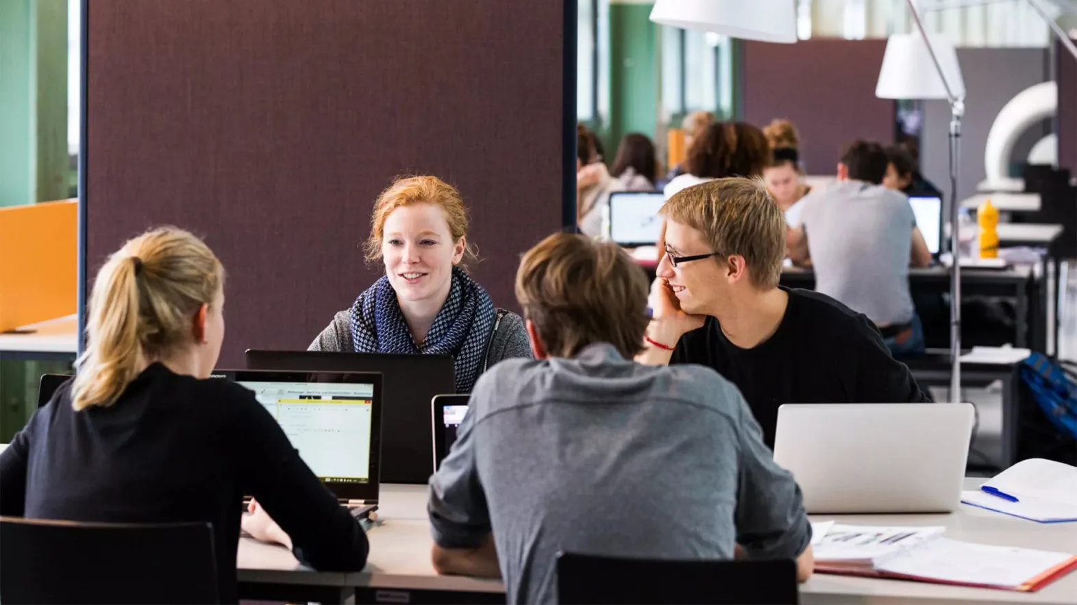 Eine Gruppe Studierender sitzt mit Laptops an einem Tisch in einem Lernraum und unterhält sich.