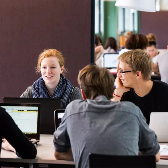 Eine Gruppe Studierender sitzt mit Laptops an einem Tisch in einem Lernraum und unterhält sich.