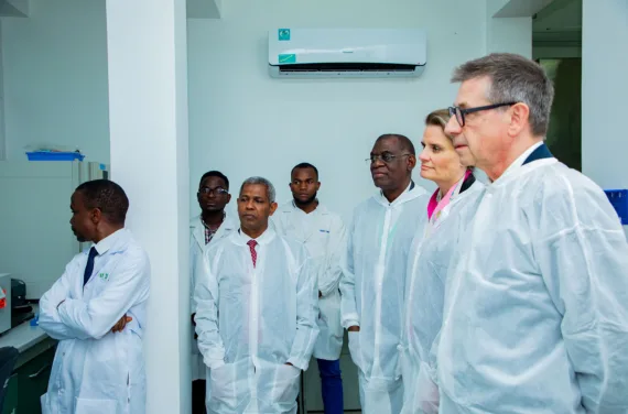 A group of people in white protective suits are standing around in a white room and listening attentively.