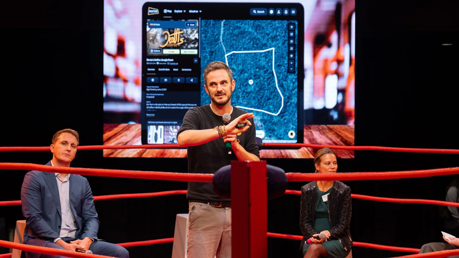 A man gestures in a red boxing ring in front of a screen showing an app interface. A man and a woman sit on gray stools in the ring behind the presenter.