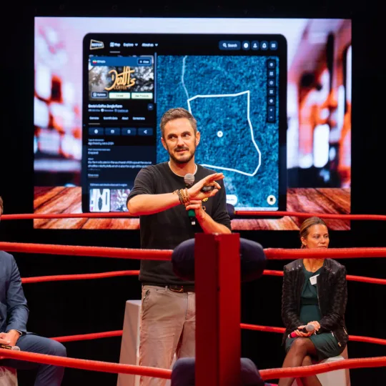 A man gestures in a red boxing ring in front of a screen showing an app interface. A man and a woman sit on gray stools in the ring behind the presenter.