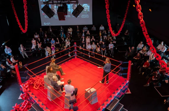 In a red boxing ring shown from above, a person talks to an audience against a black backdrop with four people sat behind them on gray stools.