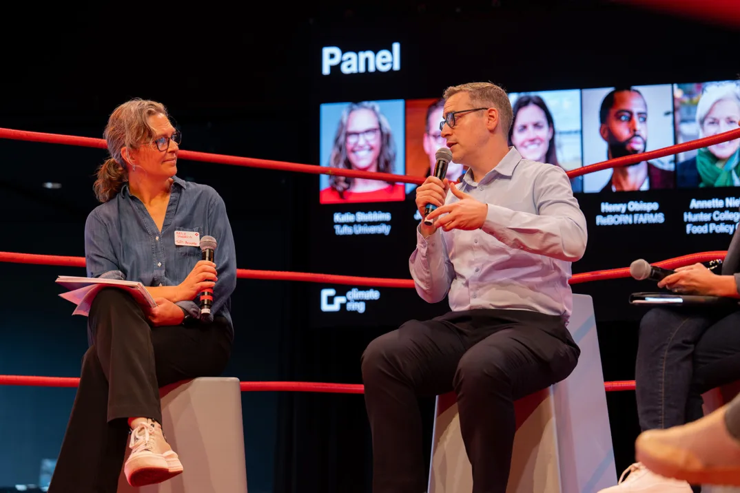 A man on a gray stool talks into a microphone. Next to him is a woman with a microphone, in the background is a screen with photos of the panelists on them.