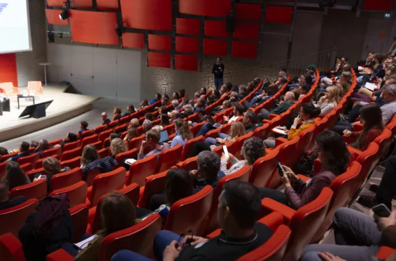 Aufnahme vom Auditorium, rund 160 Besucher sitzen auf roten Stühlen und schauen zur Bühne.