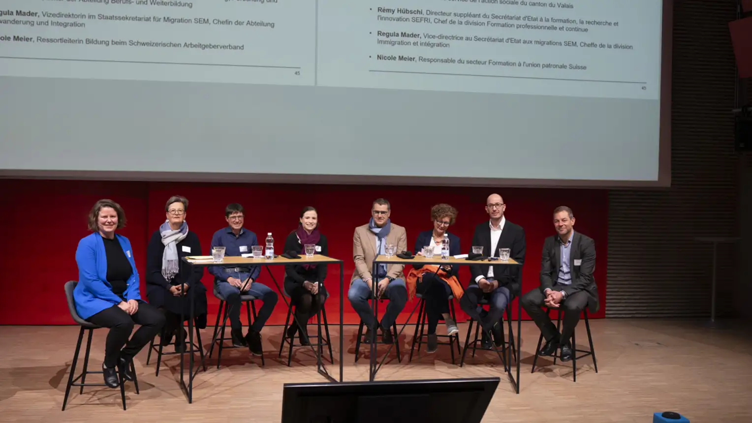 Huit personnes sont assises à deux longues tables sur la scène, elles regardent partiellement la caméra et discutent entre elles.