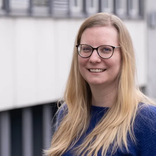 Priska Barghouti ist eine Frau mit blonden Haaren. Sie trägt eine Brille und einen violetten Pullover.