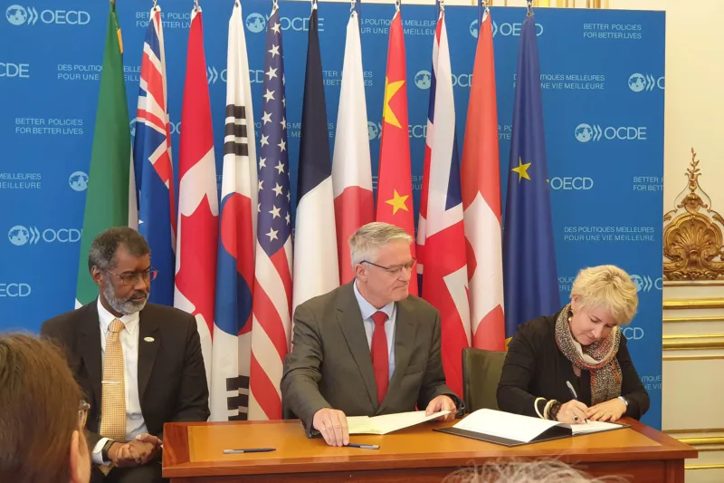 Trois personnes signent un contrat devant une collection de drapeaux.