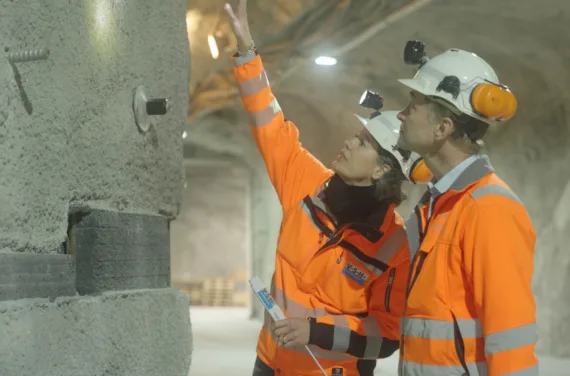 Zwei Personen in orangem Schutzanzug mit Helm stehen in einem Tunnel