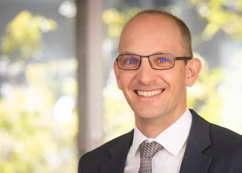 Ein Mann mit Brille und in Anzug, im Hintergrund ist ein Baum zu sehen