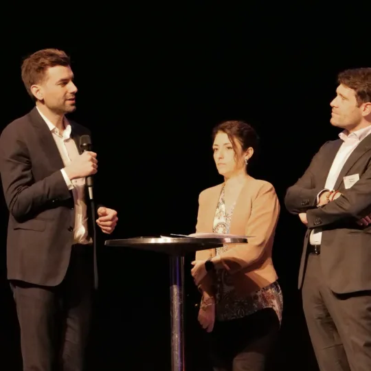 Two men and a woman are standing around a round table talking together.
