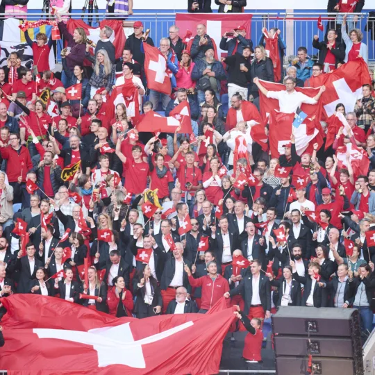 Eine grosse Gruppe an Menschen stehen auf einer Tribüne und schwenken Schweizer Fahnen.