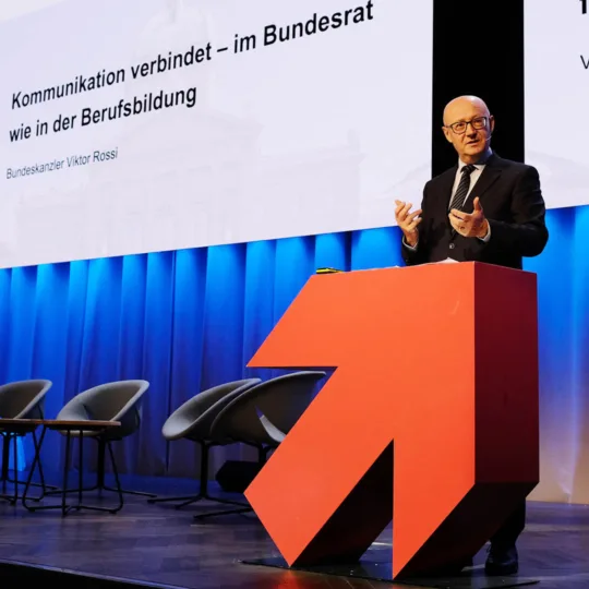 Un homme portant des lunettes et chauve prononce un discours derrière un pupitre en forme de flèche rouge.