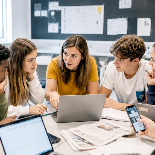 Gruppe von jungen Menschen sitzen im Halbkreis an einem Tisch, diskutierend, Laptops und Papier auf dem Tisch