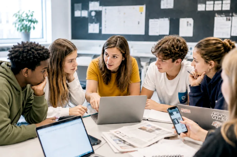 Gruppe von jungen Menschen sitzen im Halbkreis an einem Tisch, diskutierend, Laptops und Papier auf dem Tisch