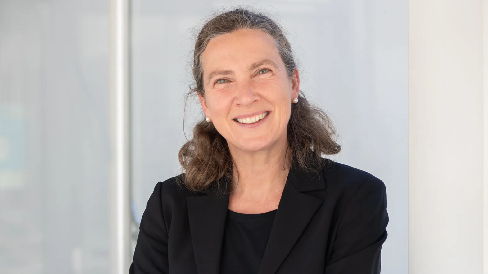 Portrait of a woman with curly hair tied back in a black blazer in front of a light background.