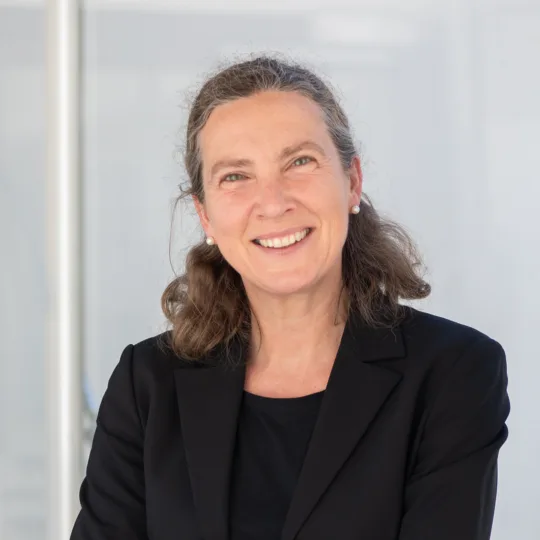 Portrait of a woman with curly hair tied back in a black blazer in front of a light background.