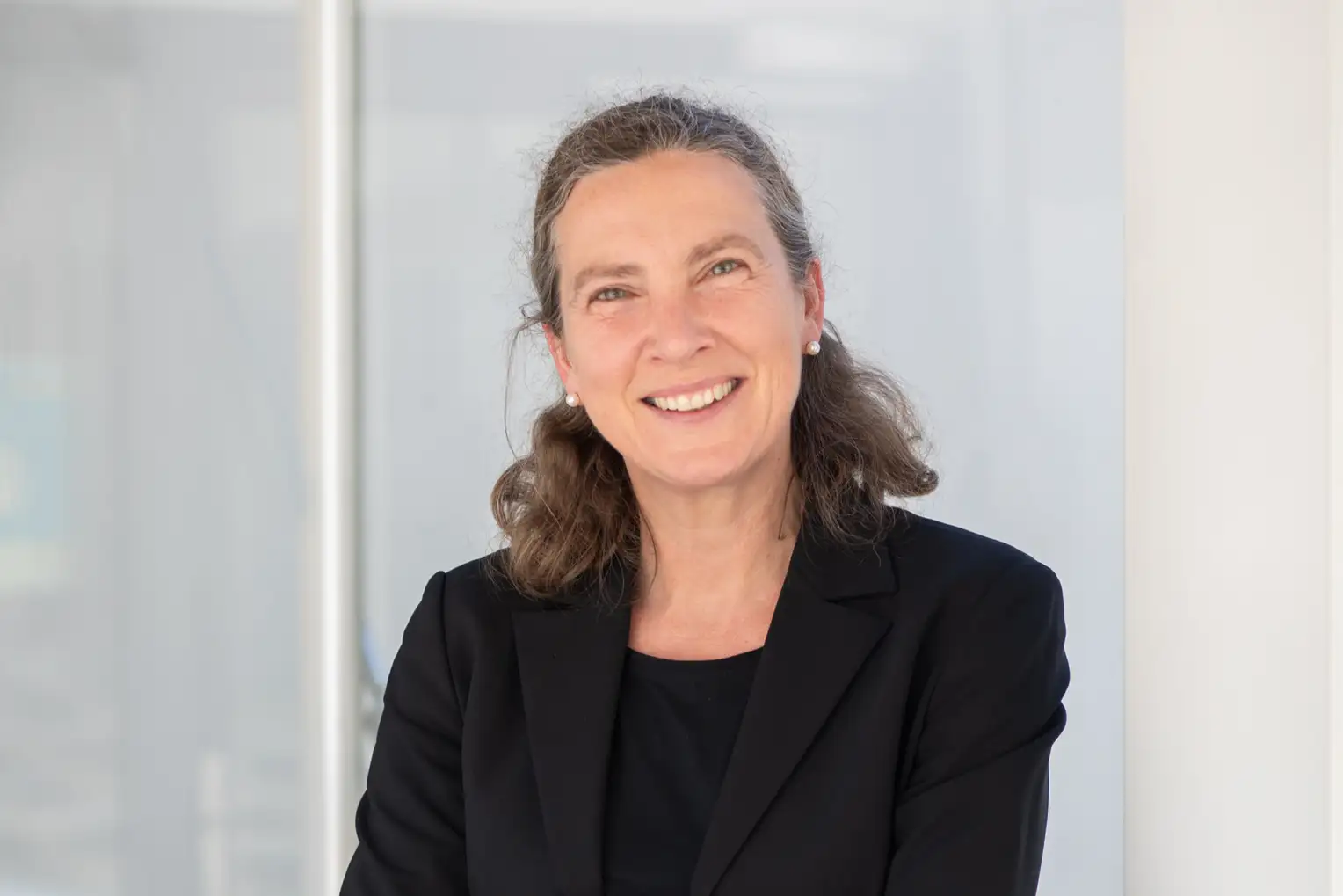 Portrait of a woman with curly hair tied back in a black blazer in front of a light background.